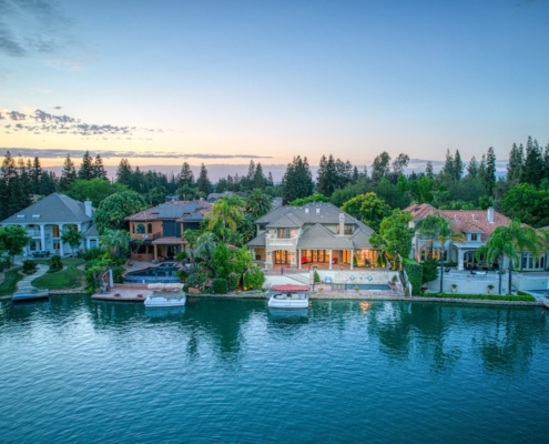 A stunning waterfront home with boats docked in the water, surrounded by lush greenery and a clear blue sky.
